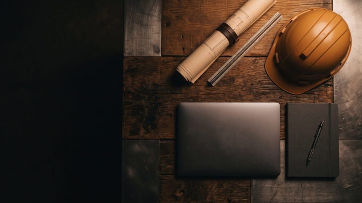 Rolled blueprints, a hard hat, a closed laptop and a notebook on a dark wooden planning table