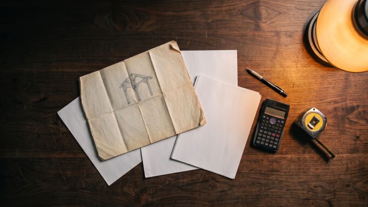 A folded structural drawing, blank sheets, a calculator, pen and tape measure on a dark desk under a warm lamp