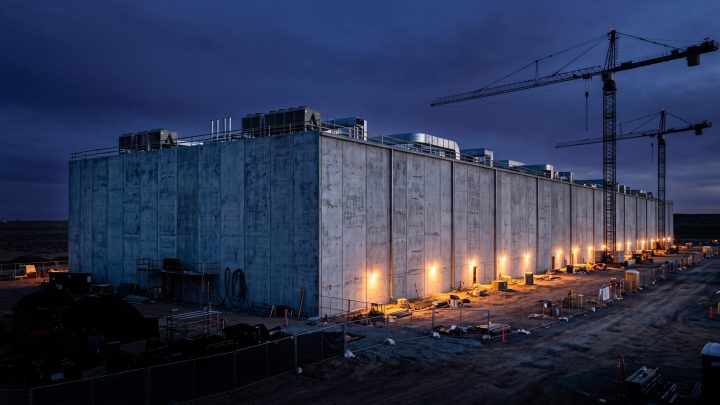 A long windowless data center under construction at dusk, with tower cranes behind it and lit work areas along its base